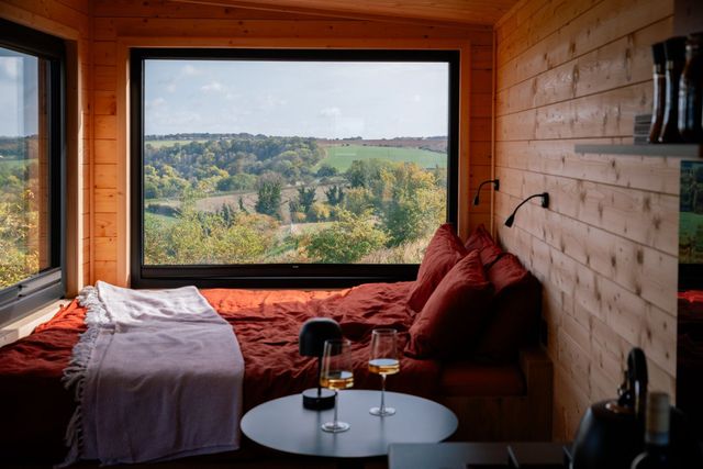 A bed by a big picture window looking out over rolling English countryside, two glasses of wine on the table